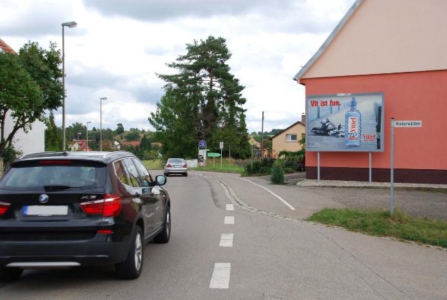 Two-lane street in a small town with a black SUV in the foreground, a red building with a billboard on the right, and parked cars along the road.