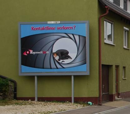 Billboard on a green wall reading 'Kontaklinsen verloren?' with a spy-style image of a person crawling into a camera lens.