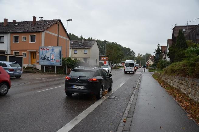 Wet street in a residential area with cars driving in both directions, a sidewalk on the right, a billboard on a building, and an overcast sky. A cyclist is visible in the distance.