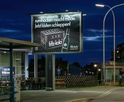 Nighttime outdoor scene with a large illuminated Fritz-kola billboard showing a bottle and crate, mounted on a tall post near a bus stop.